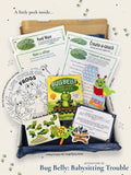 A children's activity kit labeled 'Bug Belly: Babysitting Trouble' arranged in a cardboard box with a blue fabric liner. Visible contents include printed worksheets, a coloring page, a book, a magnifying glass, and small frog-themed accessories.